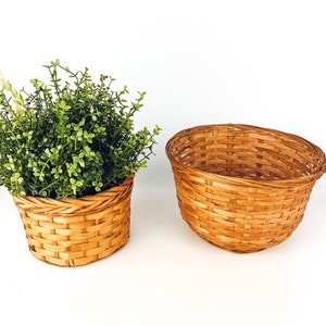 May include: Two woven baskets made of natural brown wicker. The smaller basket is filled with a green artificial plant. Both baskets are sitting on a white surface.