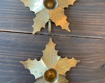 May include: Two gold metal candle holders shaped like maple leaves. Each holder has a round candle cup and a hook on the back for hanging.