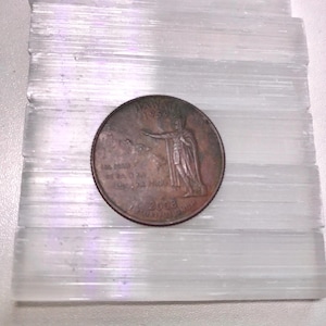 May include: A close-up of a 2003 Hawaiian state quarter on a white selenite crystal slab. The coin features a depiction of the goddess  Haumea, the Hawaiian goddess of childbirth and fertility.
