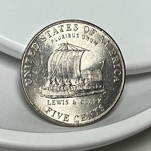 May include: A silver US five-cent coin with the inscription "United States of America" and "E Pluribus Unum" around the edge. The coin features a depiction of a boat with the text "Lewis & Clark" and "Five Cents" below.