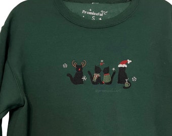 May include: Green Christmas sweatshirt with an embroidered design of four black cats wearing festive attire, including a reindeer hat, a scarf, a Santa hat, and a mug. The text "Promiseful Art" is embroidered on the top of the sweatshirt.