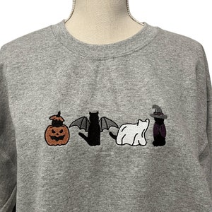 May include: A gray sweatshirt with an embroidered design of a pumpkin, a bat, a ghost, and a witch. The design is in black, white, orange, and purple.