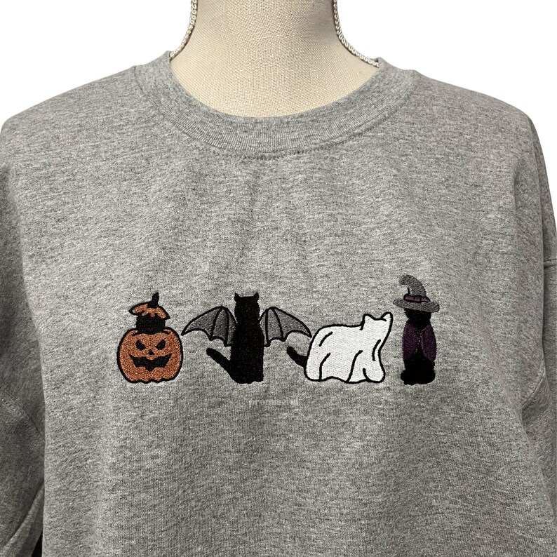 May include: A gray sweatshirt with an embroidered design of a pumpkin, a bat, a ghost, and a witch. The design is in black, white, orange, and purple.