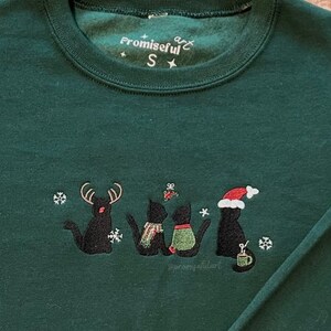May include: Green Christmas jumper with black embroidered cats wearing Santa hats and reindeer antlers. The jumper has the text "Promiseful" and a star on the front.