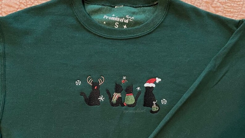 May include: Green Christmas jumper with black embroidered cats wearing Santa hats and reindeer antlers. The jumper has the text "Promiseful" and a star on the front.