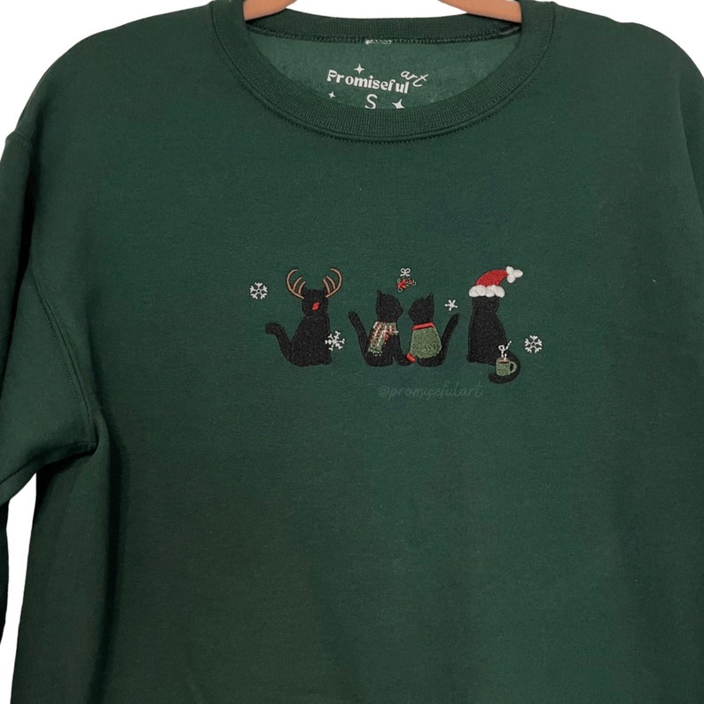 May include: Green Christmas sweatshirt with an embroidered design of four black cats wearing festive attire, including a reindeer hat, a scarf, a Santa hat, and a mug. The text "Promiseful Art" is embroidered on the top of the sweatshirt.