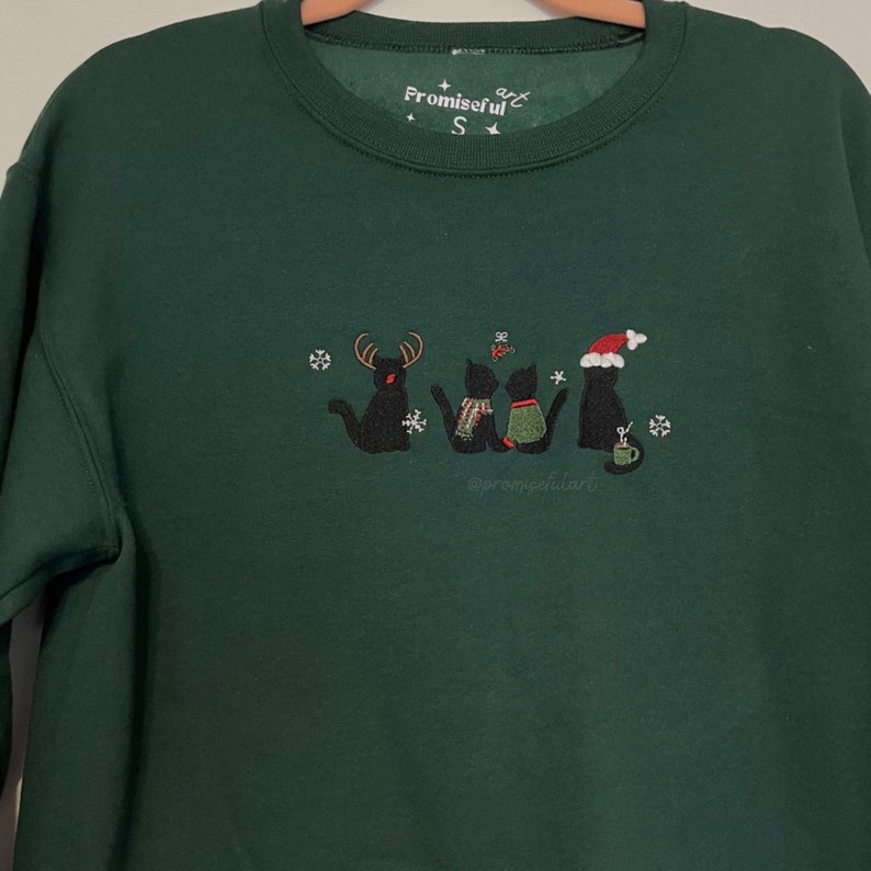May include: A dark green sweatshirt with an embroidered design of four black cats, one wearing a Santa hat, another with antlers, and two with scarves. The text "Promiseful Art" is embroidered on the top of the sweatshirt.