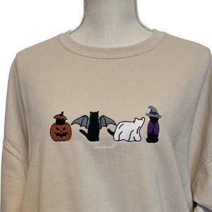 May include: A beige sweatshirt with an embroidered design of four cats dressed up for Halloween. The cats are dressed as a pumpkin, a bat, a ghost, and a witch.