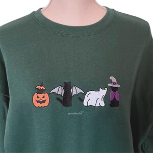 May include: A dark green sweatshirt with an embroidered design of a pumpkin, a bat, a ghost, and a witch. The design is in black, white, and purple.