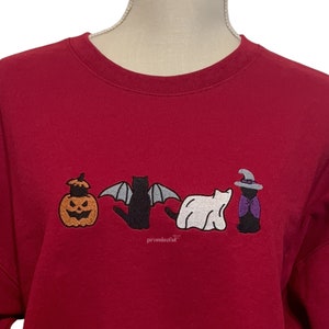 May include: A red sweatshirt with an embroidered design of a pumpkin, bat, ghost, and witch. The design is in black, white, and purple.