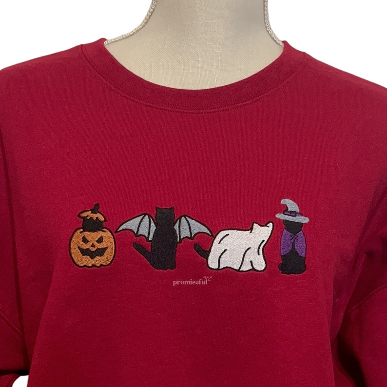 May include: A red sweatshirt with an embroidered design of a pumpkin, bat, ghost, and witch. The design is in black, white, and purple.