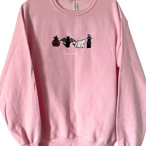 May include: A pink sweatshirt with an embroidered design of a pumpkin, a bat, a ghost, and a witch. The text "promissful" is embroidered below the design.