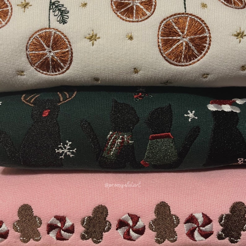 May include: Three stacked sweatshirts with embroidered festive designs. The top sweatshirt is white with orange citrus fruit and gold stars. The middle sweatshirt is green with black cats and a reindeer. The bottom sweatshirt is pink with gingerbread men and candy canes.