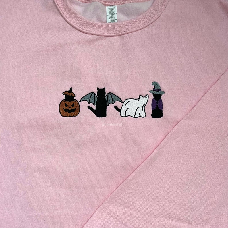 May include: A pink sweatshirt with an embroidered design of a pumpkin, a bat, a ghost, and a witch cat. The design is in black, white, orange, and purple.
