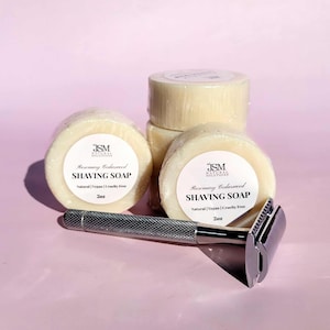 Peut inclure: Plusieurs savons à raser et un rasoir de sécurité sur une surface rose. Les savons ronds sont emballés dans du plastique transparent et étiquetés "Rosemary Cedarwood Shaving Soap". Le rasoir est de couleur argentée.