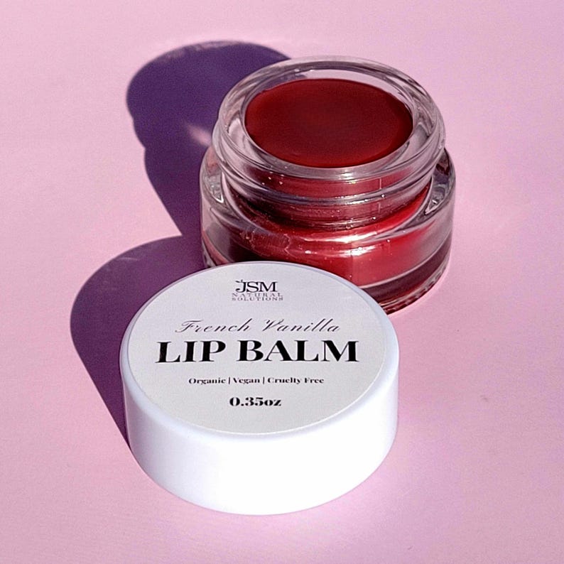 May include: A jar of French Vanilla lip balm with a white lid and clear glass jar. The lip balm is a deep red color. The lid has the text "French Vanilla LIP BALM" and "Organic | Vegan | Cruelty Free 0.35oz".