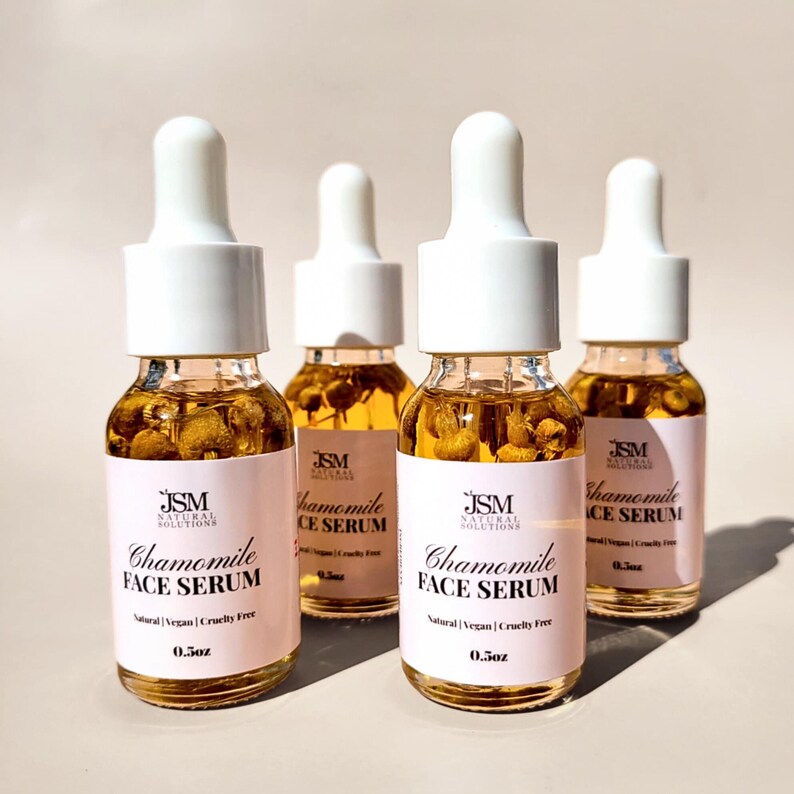 May include: Four glass bottles of Chamomile Face Serum with white droppers. Each bottle has a pink label with the text "Chamomile FACE SERUM" and "Natural | Vegan | Cruelty Free". The bottles contain dried chamomile flowers and are 0.5oz.