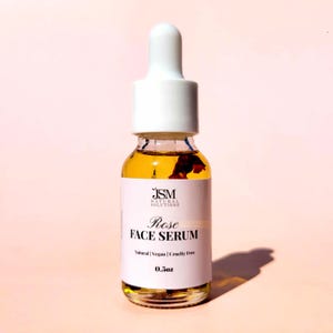 May include: A clear glass bottle of Rose Face Serum with a white dropper top. The serum is amber-colored and contains rose petals. The label reads "Rose FACE SERUM" and "Natural | Vegan | Cruelty Free". The bottle contains 0.5 oz of serum.
