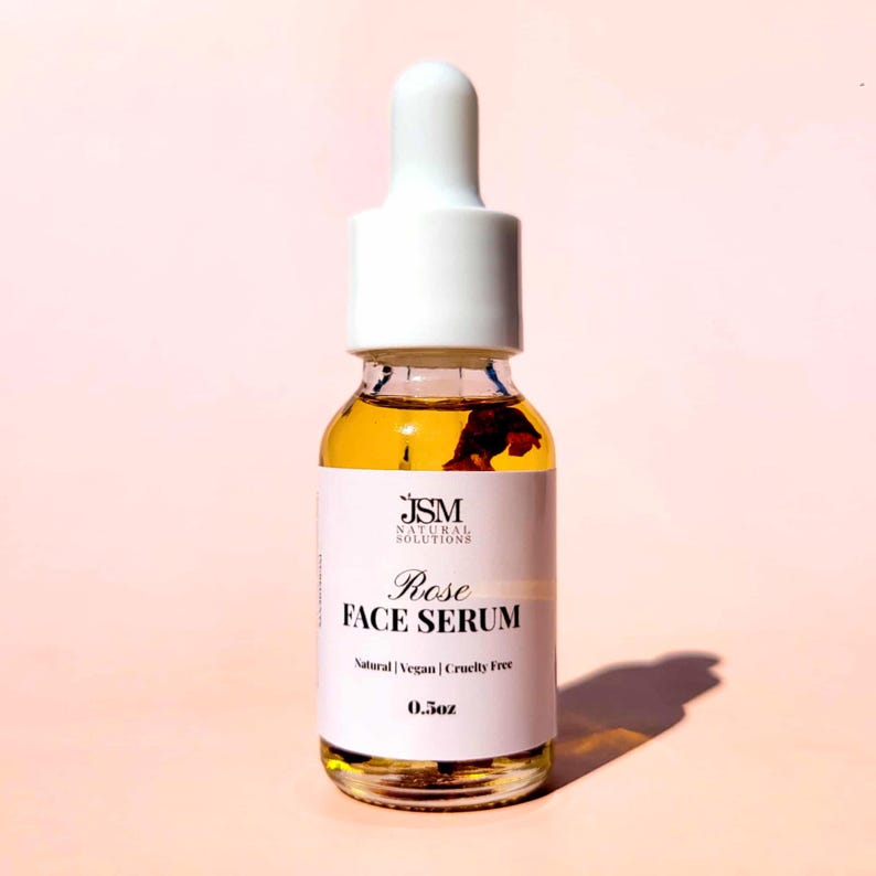 May include: A clear glass bottle of Rose Face Serum with a white dropper top. The serum is amber-colored and contains rose petals. The label reads "Rose FACE SERUM" and "Natural | Vegan | Cruelty Free". The bottle contains 0.5 oz of serum.