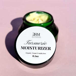 May include: A small jar of turmeric moisturiser with a green glass base and a white lid. The lid displays the text "Turmeric Moisturizer" and "0.5oz". The product is organic, vegan, and cruelty-free.