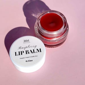 May include: A jar of raspberry lip balm with a white lid. The lid is off, revealing the red balm inside the clear glass jar. The lid has the text "Raspberry Lip Balm" and "0.35oz". The background is a light pink color.