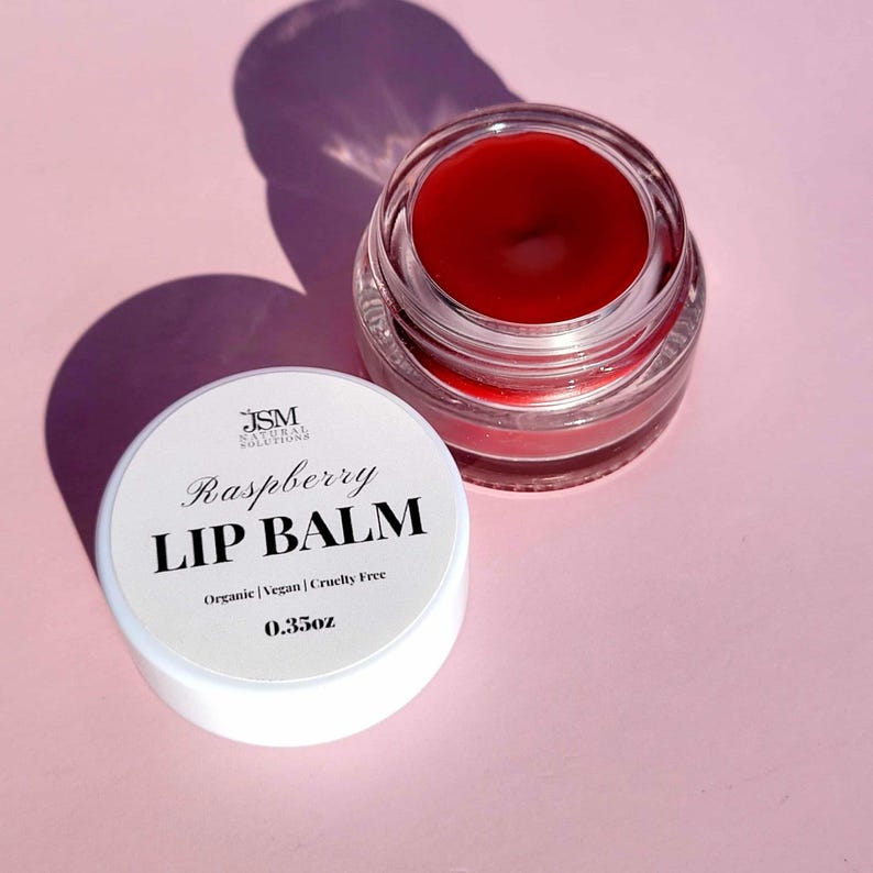 May include: A jar of raspberry lip balm with a white lid. The lid is off, revealing the red balm inside the clear glass jar. The lid has the text "Raspberry Lip Balm" and "0.35oz". The background is a light pink color.