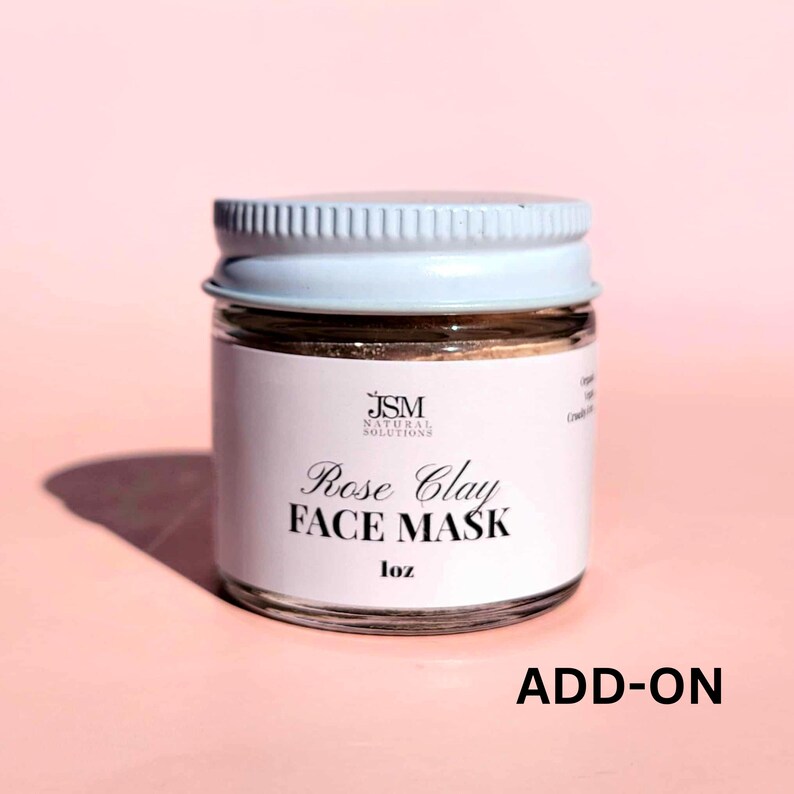 May include: A small glass jar with a white lid and a label that reads "Rose Clay FACE MASK" in elegant script. The label also includes the text "JSM NATURAL SOLUTIONS" and "1 oz". The jar is filled with a brown substance.