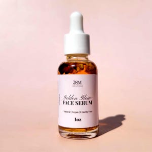 May include: A clear glass bottle of Golden Glow Face Serum with a white dropper top. The bottle contains a golden liquid with small particles. The label reads "JSM Natural Solutions" and "Natural | Vegan | Cruelty Free". The bottle is 1 oz.