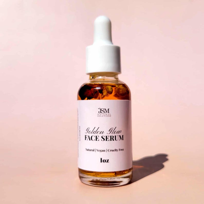 May include: A clear glass bottle of Golden Glow Face Serum with a white dropper top. The bottle contains a golden liquid with small particles. The label reads "JSM Natural Solutions" and "Natural | Vegan | Cruelty Free". The bottle is 1 oz.