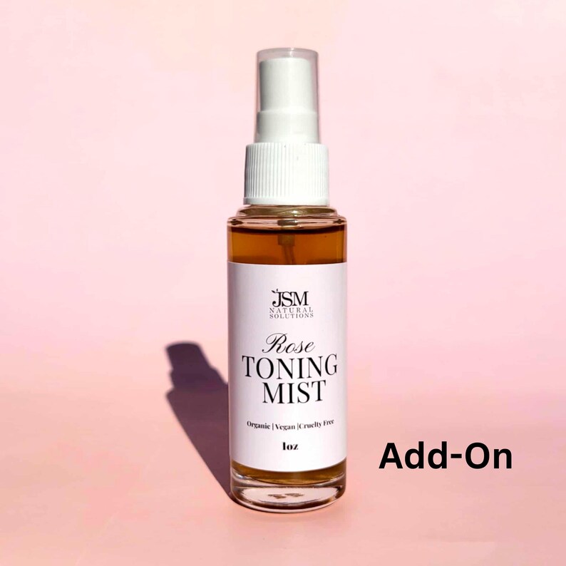 May include: A clear glass spray bottle filled with a light brown liquid, labeled "Rose Toning Mist." The label includes the text "JSM Natural Solutions," and "Organic | Vegan | Cruelty Free." The bottle is 1 oz and has a white spray nozzle. The background is a soft pink.