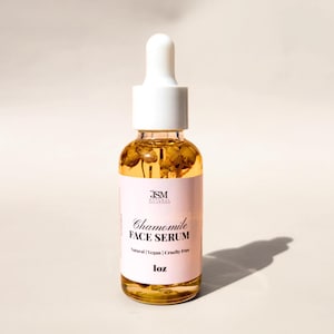 May include: A clear glass bottle of Chamomile Face Serum with a white dropper top. The serum is a golden color and contains dried chamomile flowers. The label is pink with black text that reads "Chamomile Face Serum". The bottle is 1 oz.