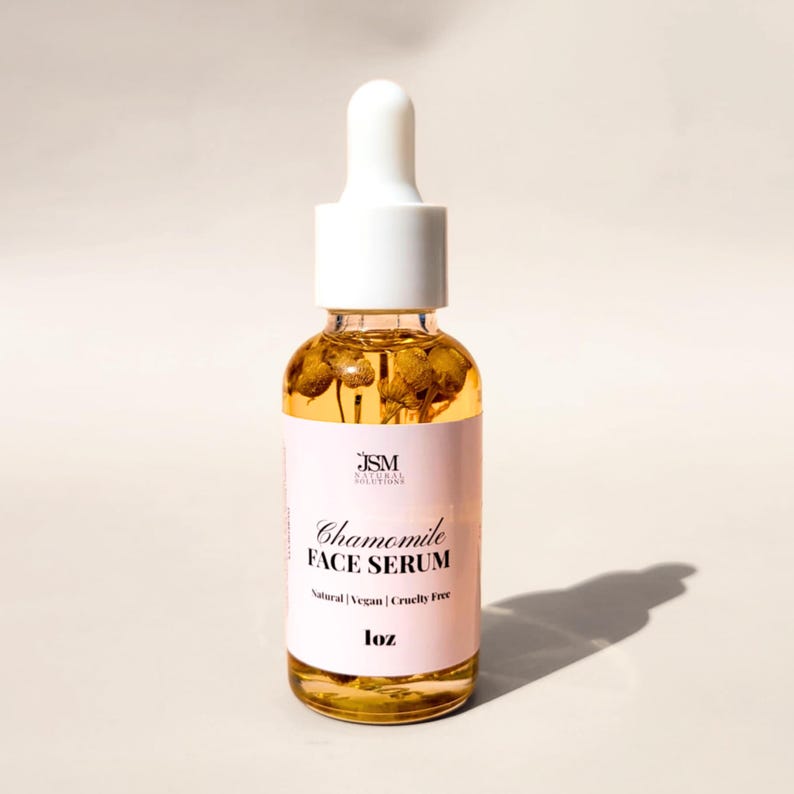 May include: A clear glass bottle of Chamomile Face Serum with a white dropper top. The serum is a golden color and contains dried chamomile flowers. The label is pink with black text that reads "Chamomile Face Serum". The bottle is 1 oz.