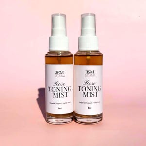 May include: Two clear glass spray bottles filled with a brown liquid, labeled "Rose Toning Mist." The bottles have white labels with black text and a white spray nozzle. The background is a soft pink.