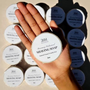 May include: A hand holding a round shaving soap with the text "Rosemary Cedarwood SHAVING SOAP". The soap is labeled "Natural | Vegan | Cruelty Free" and is 57g. Several other soaps are visible in the background.