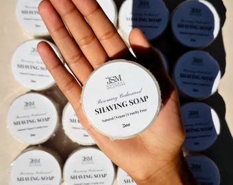 May include: A hand holding a round shaving soap with the text "Rosemary Cedarwood SHAVING SOAP". The soap is labeled "Natural | Vegan | Cruelty Free" and is 2oz. Several other soaps are visible in the background.
