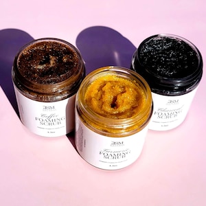 May include: Three glass jars of foaming scrub on a pink surface. The jars contain coffee, turmeric, and charcoal scrubs. Each jar has a white label with black text. The coffee scrub is dark brown, the turmeric scrub is yellow, and the charcoal scrub is black.