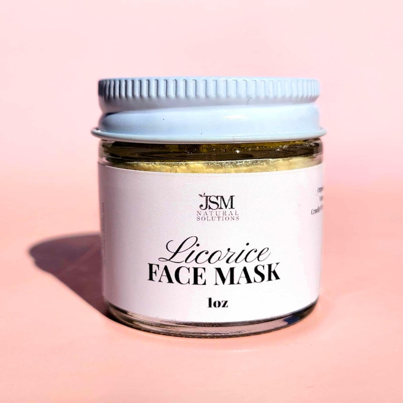 May include: A small glass jar of licorice face mask with a light blue lid. The jar has a white label with the text "Licorice FACE MASK" and "1 oz". The background is a solid pink.