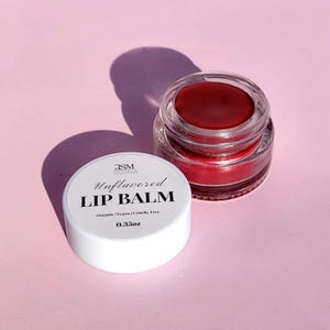 May include: An open, clear glass jar of red lip balm next to a white lid. The lid is labeled "Unflavored LIP BALM" with "Organic | Vegan | Cruelty Free" and "0.35oz" printed below. The background is a light pink.