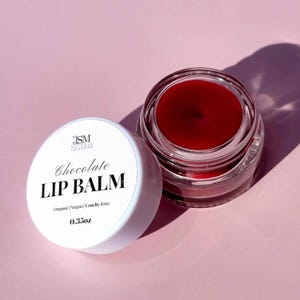 May include: A jar of chocolate lip balm with a white lid, sitting next to an open jar of the balm. The balm is a deep red color. The lid has the text "Chocolate LIP BALM" and "0.35oz". The product is labeled as organic, vegan, and cruelty-free.