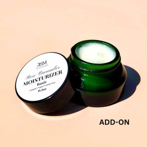 May include: An open, dark green glass jar of moisturizer with a white cream inside, next to its lid. The lid is white with black text that reads "Rose Cucumber Moisturizer Basic" and "0.5oz". The jar is on a light pink surface.