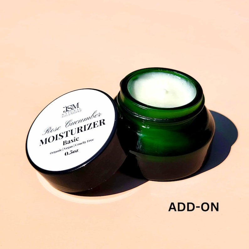 May include: An open, dark green glass jar of moisturizer with a white cream inside, next to its lid. The lid is white with black text that reads "Rose Cucumber Moisturizer Basic" and "0.5oz". The jar is on a light pink surface.