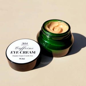 Könnte beinhalten: Ein offenes, grünes Glas mit Augencreme neben dem Deckel. Die Creme hat eine hellpfirsichfarbene Farbe und ein Wirbeldesign. Der Deckel ist schwarz mit einem weißen Etikett mit der Aufschrift "Caffeine EYE CREAM" und "0,5 oz".