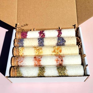 Peut inclure: Coffret cadeau contenant six tubes en verre remplis de sels de bain blancs et de diverses fleurs et herbes séchées. Les tubes sont scellés avec des bouchons en liège et disposés dans une boîte blanche avec un couvercle en carton brun.