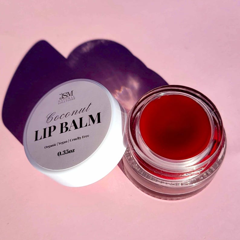 May include: A jar of red lip balm next to its white lid. The lid has the text "Coconut LIP BALM" and "Organic | Vegan | Cruelty Free" and "0.35oz". The lip balm is in a clear glass jar. The background is pink.