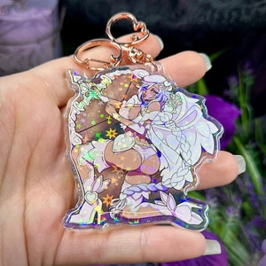 Op de afbeelding: Een heldere acryl sleutelhanger met een anime-stijl personage met pijl en boog. Het personage is in wit en bruin met holografische details. De sleutelhanger heeft een roségoudkleurige sluiting.