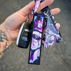 May include: A car key and keychain with a black and purple design. The keychain features a cartoon illustration of a woman with purple and white hair. The word "Devious" is printed on the keychain.