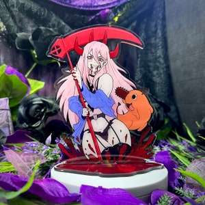 Op de afbeelding: Anime-stijl acrylfiguur met een personage met roze haar, rode hoorns en een zeis. De figuur staat op een witte basis met een rood bloedachtig ontwerp, met een oranje wezen. Omringd door paarse bloemen.