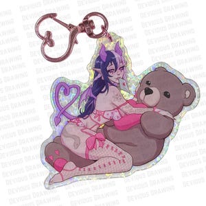 Op de afbeelding: Een holografische sleutelhanger met een cartoon demonmeisje en een teddybeer. Het meisje heeft paars haar en hoorns, draagt roze lingerie en houdt een kaart vast. De sleutelhanger heeft een hartvormige sluiting en de tekst "DEVIOUS DRAWING".