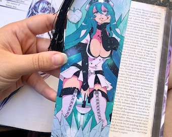 Op de afbeelding: Een bladwijzer met een manga-stijl illustratie van een vrouwelijk personage met turquoise haar, gekleed in een wit en zwart outfit. De bladwijzer heeft een zwarte kwast en ligt op een open boek.