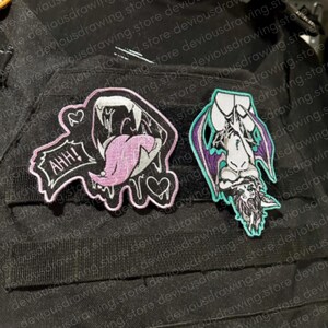 May include: Two embroidered patches on a black fabric surface. The left patch is a black shape with a pink tongue, white teeth, and the text AHH! with two hearts. The right patch is a purple and white winged creature hanging upside down.